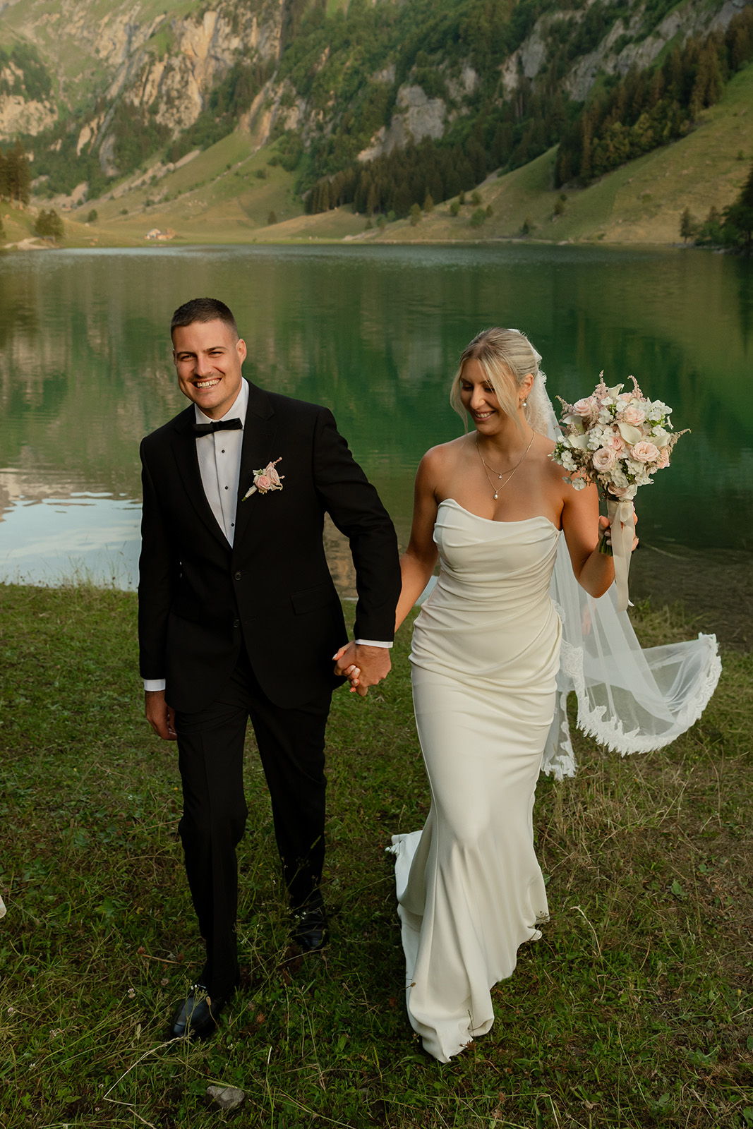Bride and groom walking by a lake, holding hands and smiling.