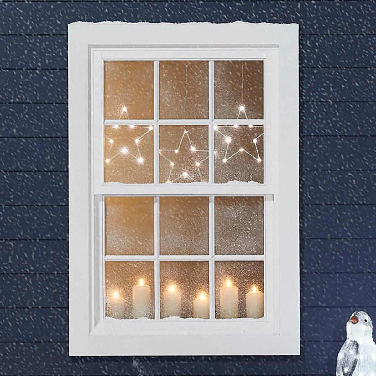 A outdoor view of a blue house at Christmas. In the window there are candles and star lights hanging above.