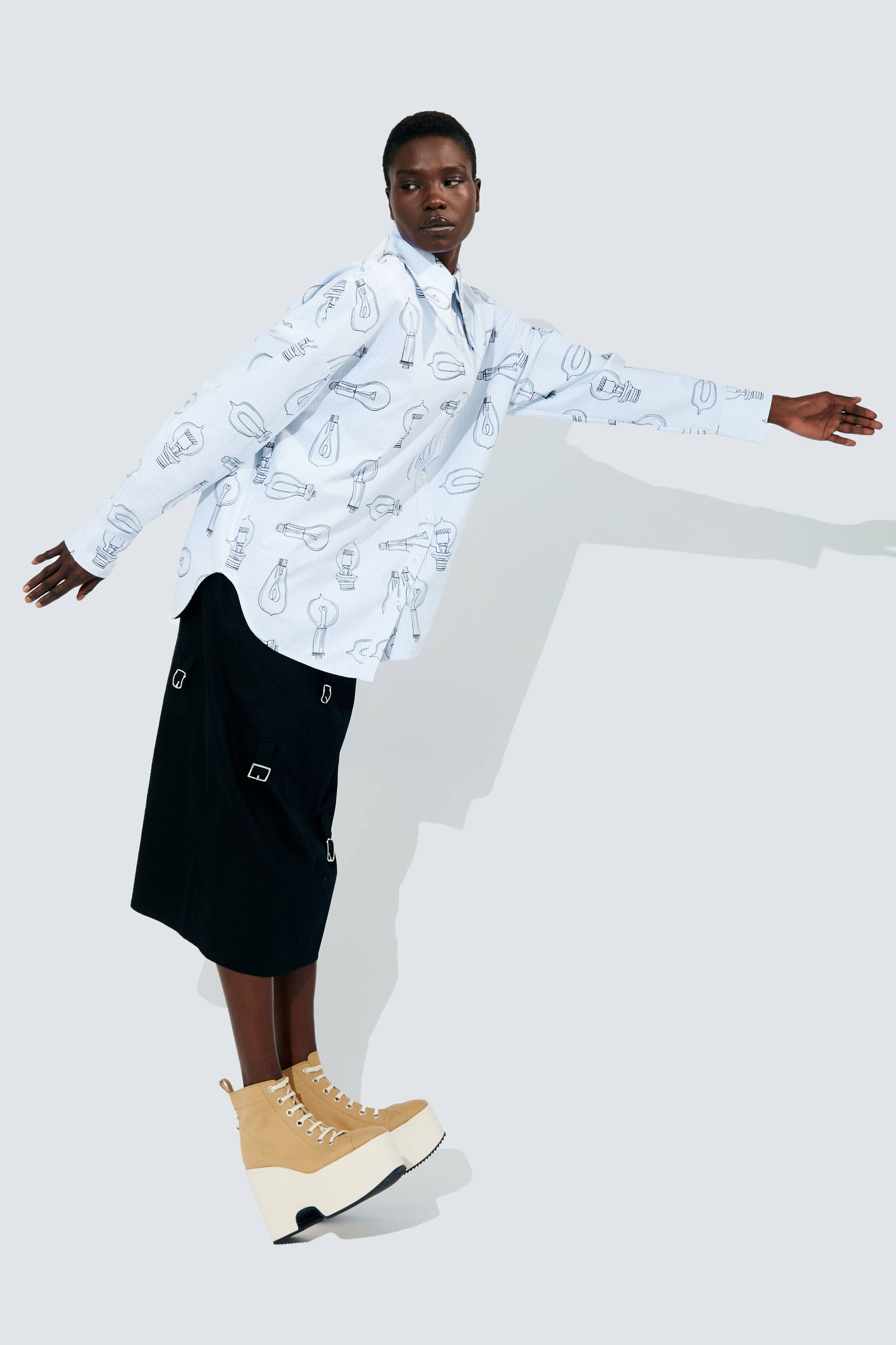 Person wearing a patterned shirt and black skirt poses against a light background.