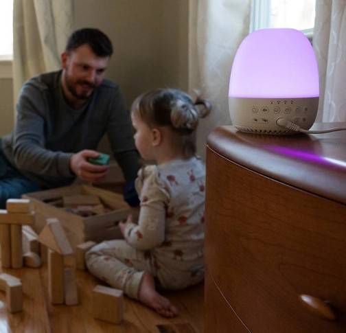 Yogasleep Light to Rise Sound Machine and Sleep Trainer Clock on drawers with toddler and dad playing in the background.