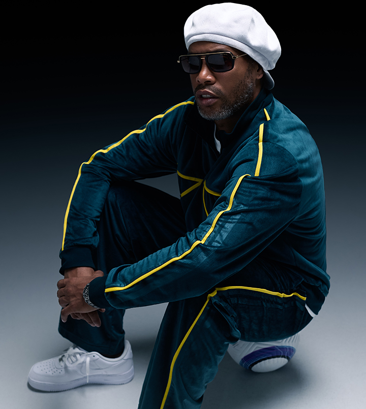 A person in a tracksuit and hat, squatting against a dark background.