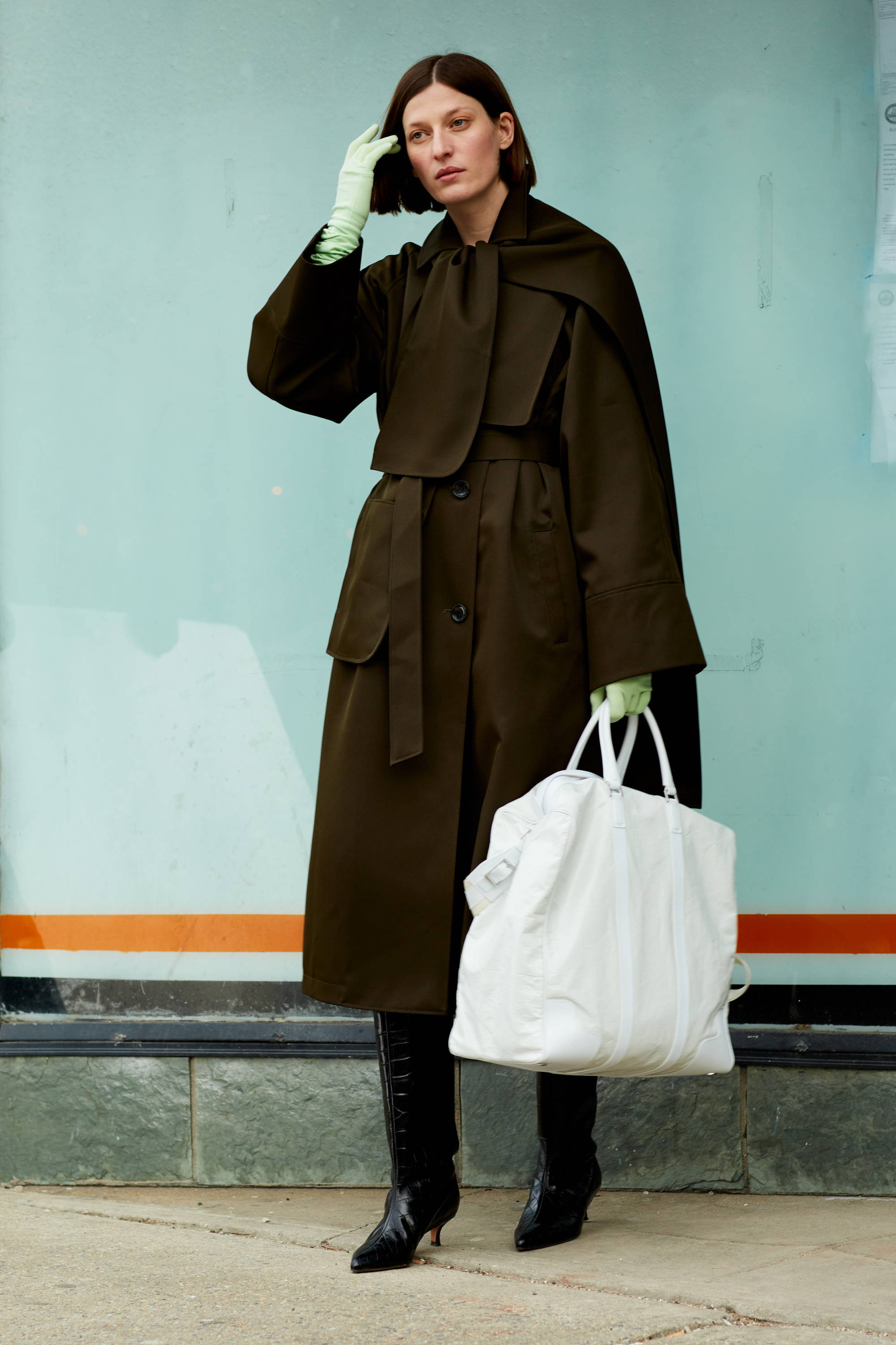 A woman in a brown coat holds a white bag against a colorful background.