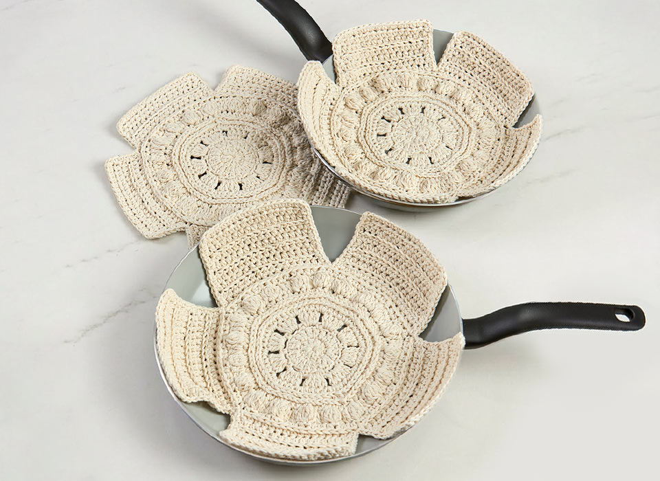 Three decorative frying pans with crocheted pot savers in light yarn.
