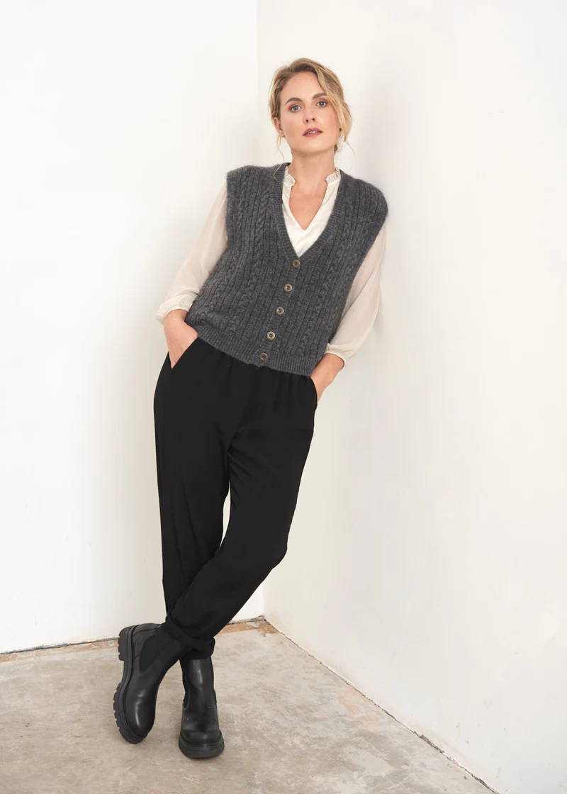A model wearing a grey sleeveless knitted waistcoat with a off white blouse underneath, black trousers and black chunky leather chelsea boots
