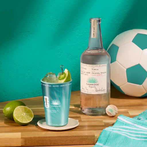 A refreshing margarita, a bottle of Casamigos tequila, limes, and a soccer ball on a wooden table.