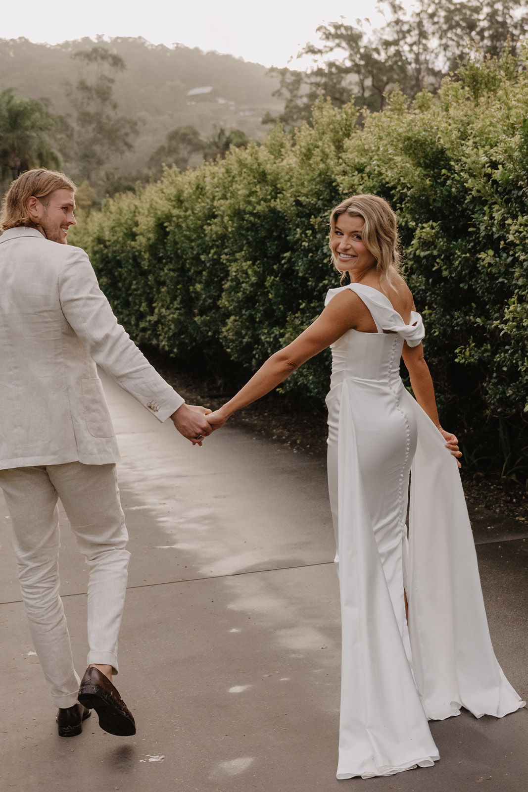 Couple holding hands, walking on a path surrounded by greenery, wearing formal attire.