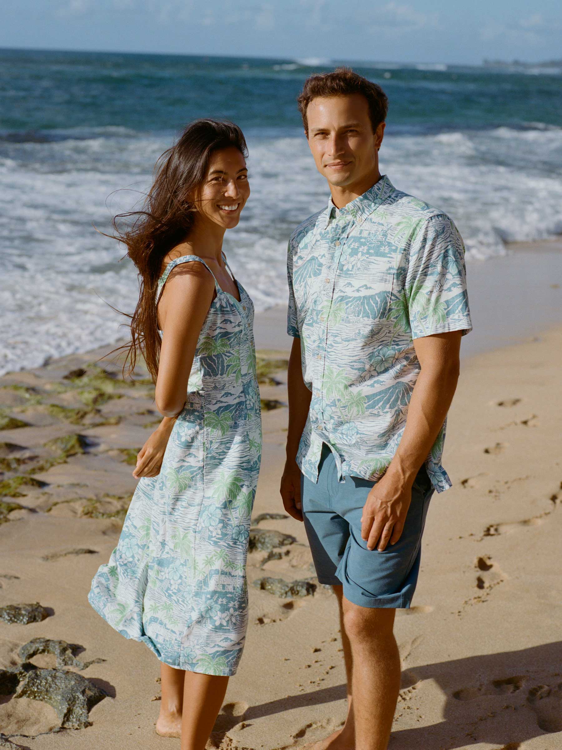 A couple in matching outfits stands on a beach by the ocean.