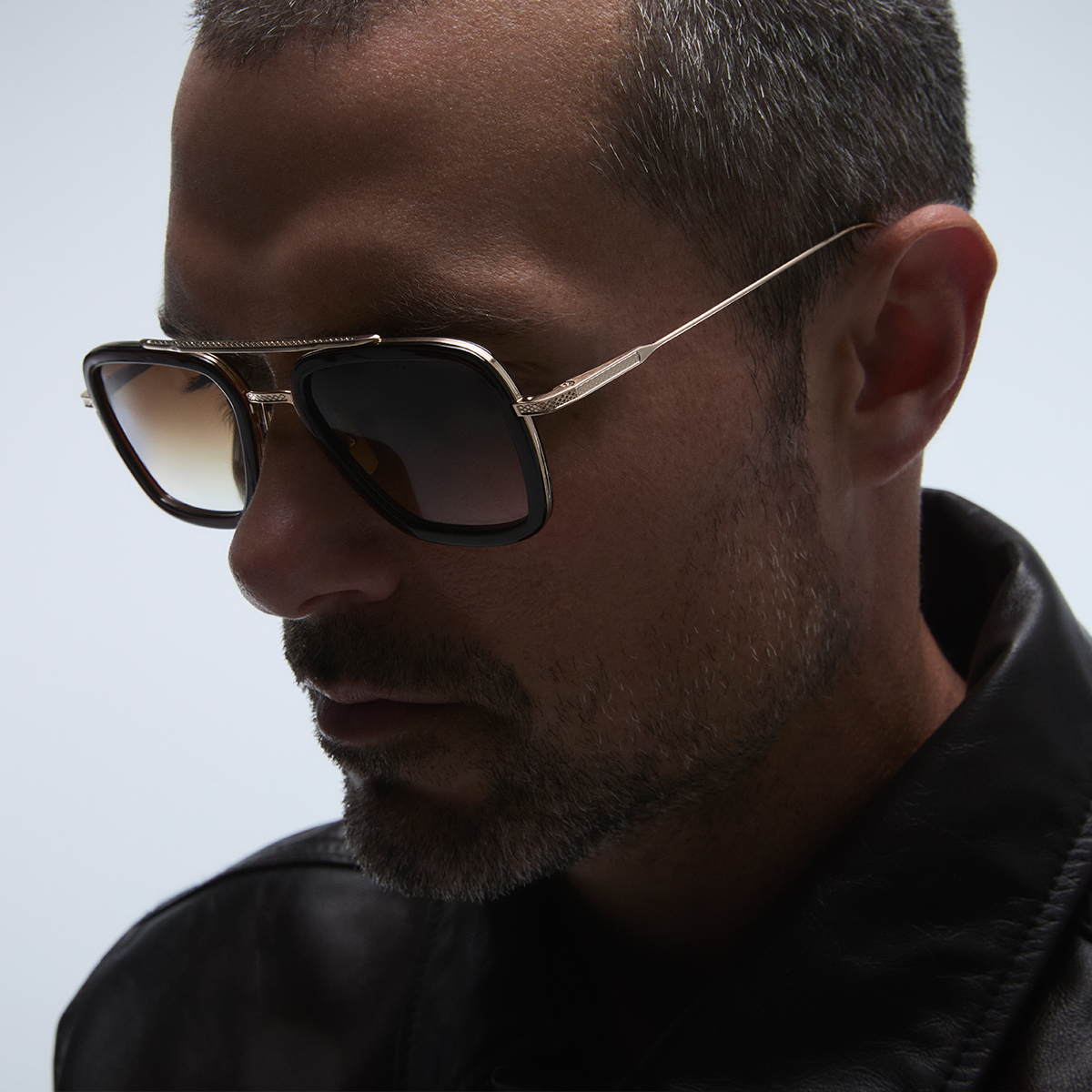 Man wearing sunglasses and a black leather jacket, looking down thoughtfully.
