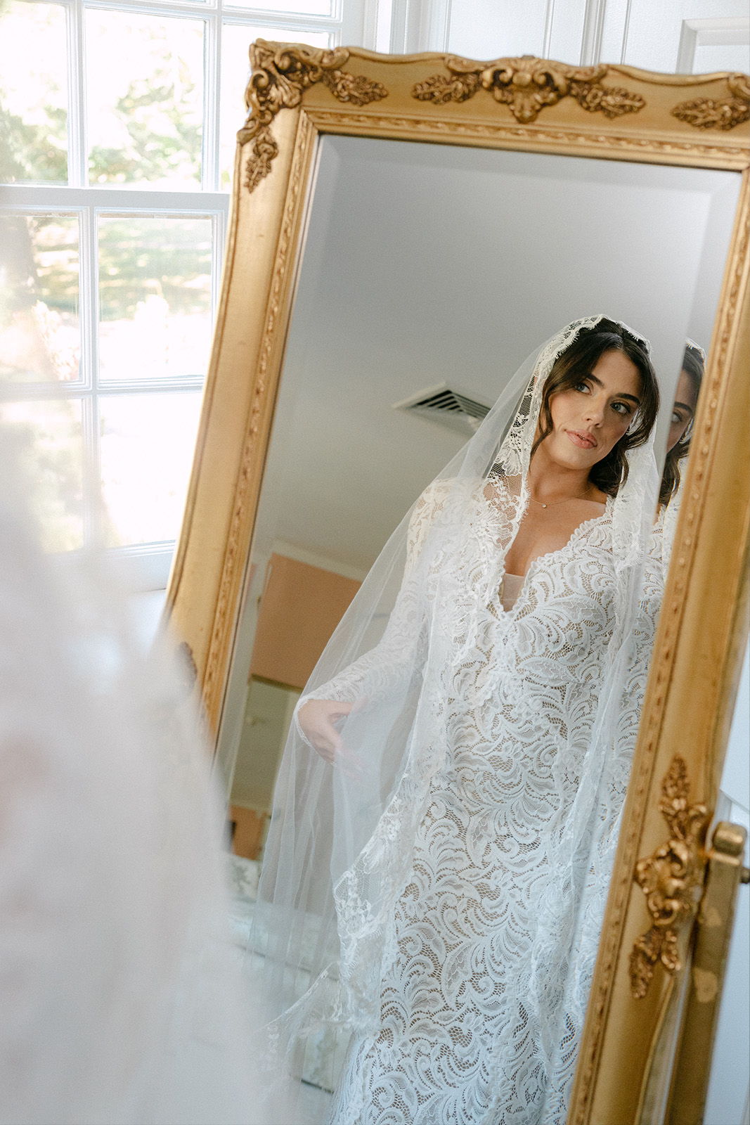 Bride in a lace wedding dress gazes at her reflection in a mirror.
