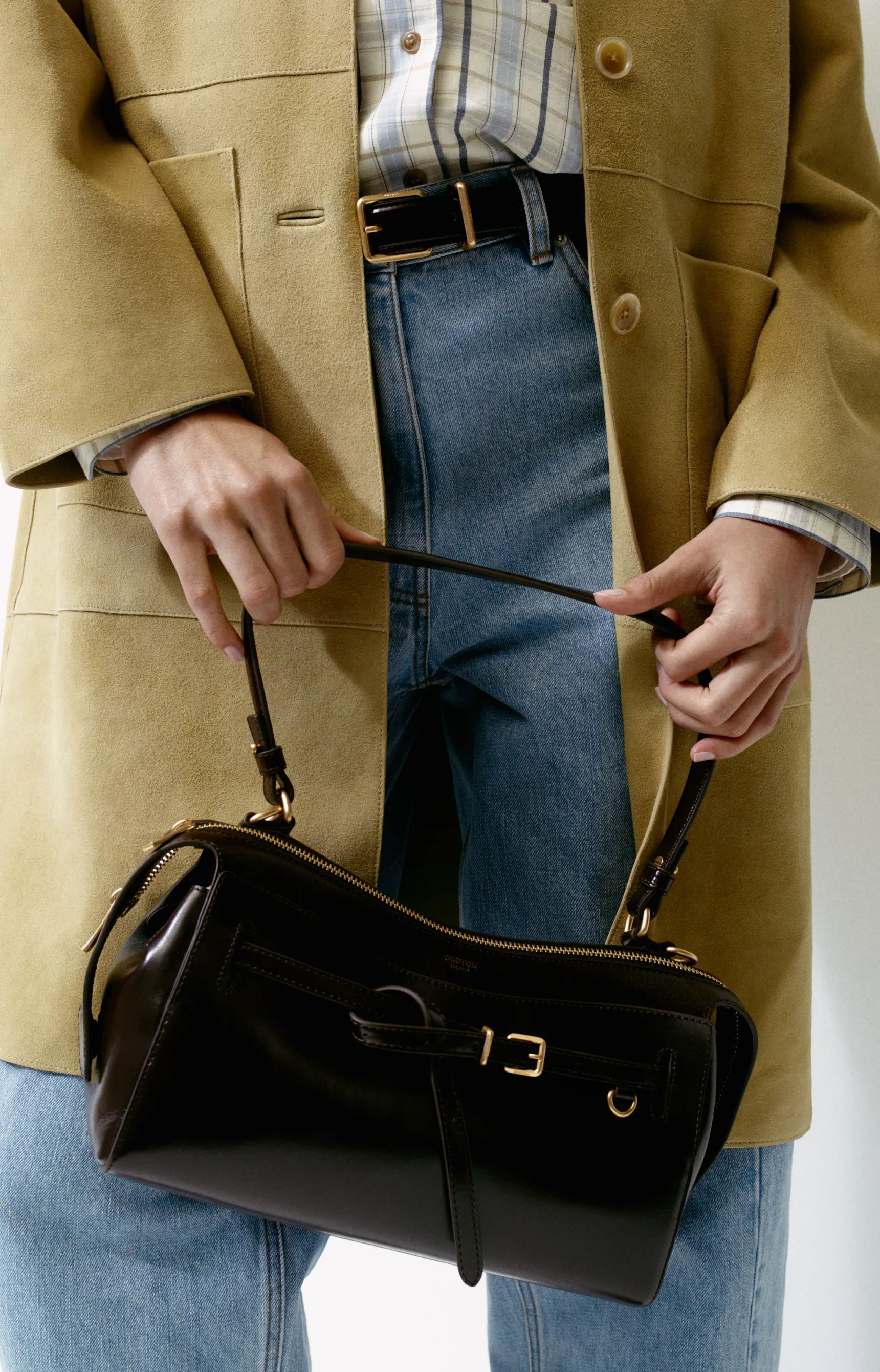 Person holding a stylish black handbag while wearing a beige coat and blue jeans.