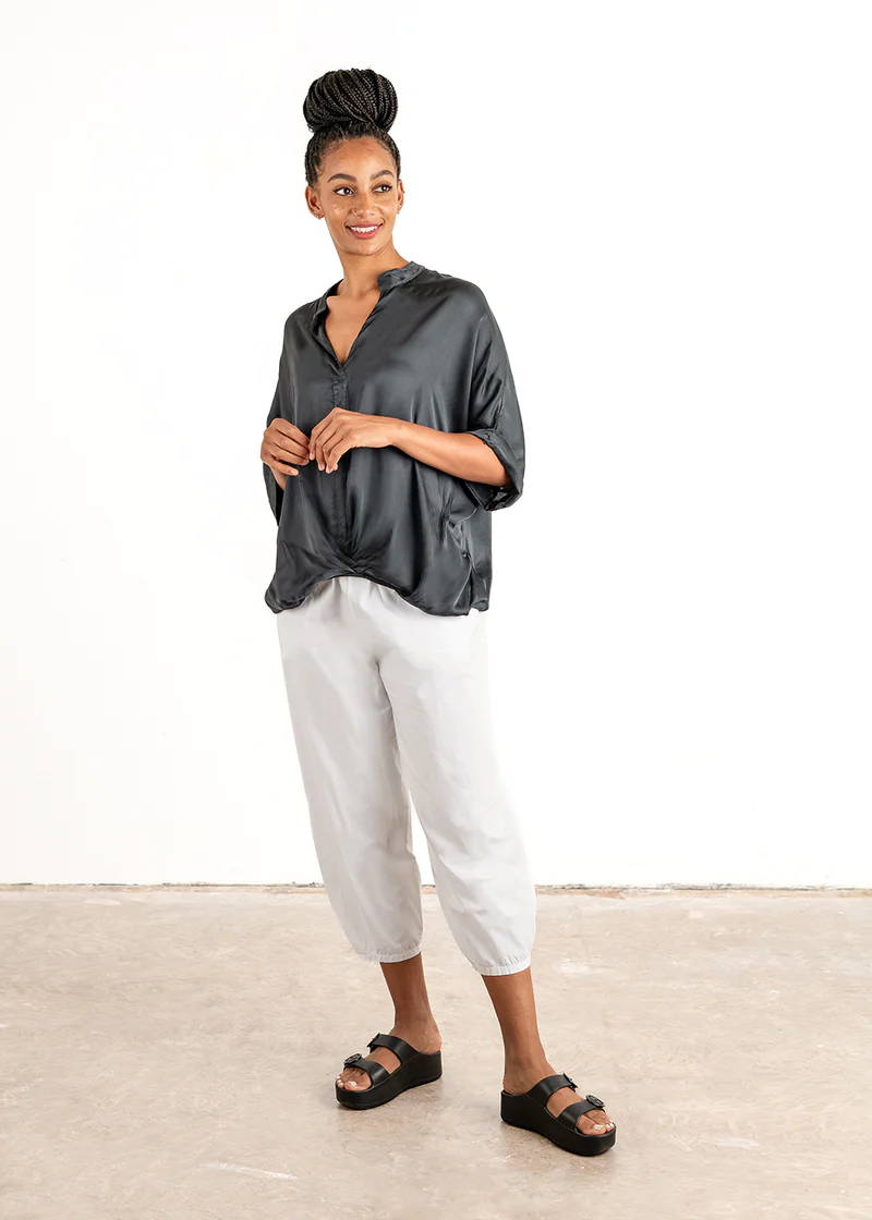 A model wearing a dark grey satin blouse with an elasticated waisr and 3/4 sleeves over white trousers and black slides
