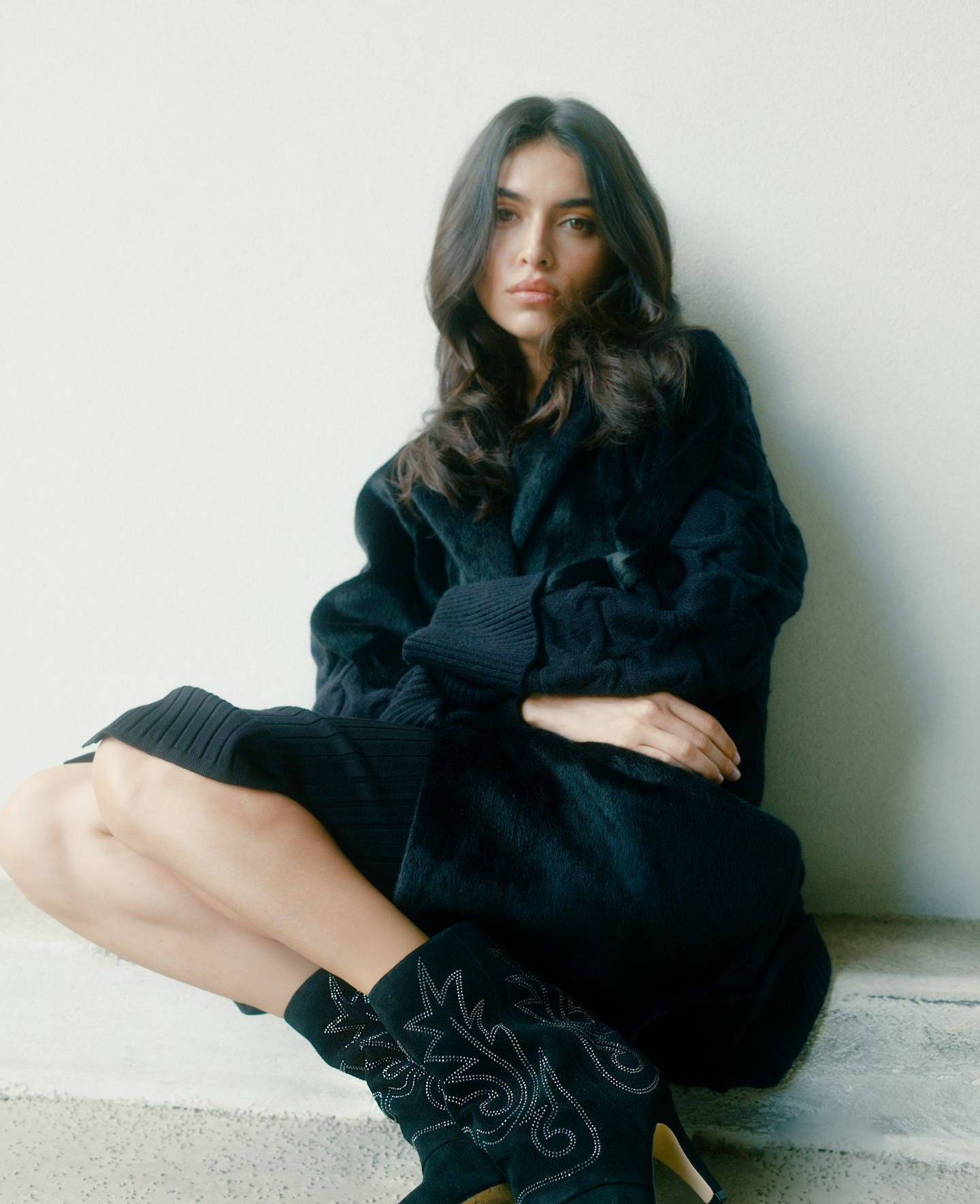 A woman in a black outfit sitting against a wall, wearing stylish boots.