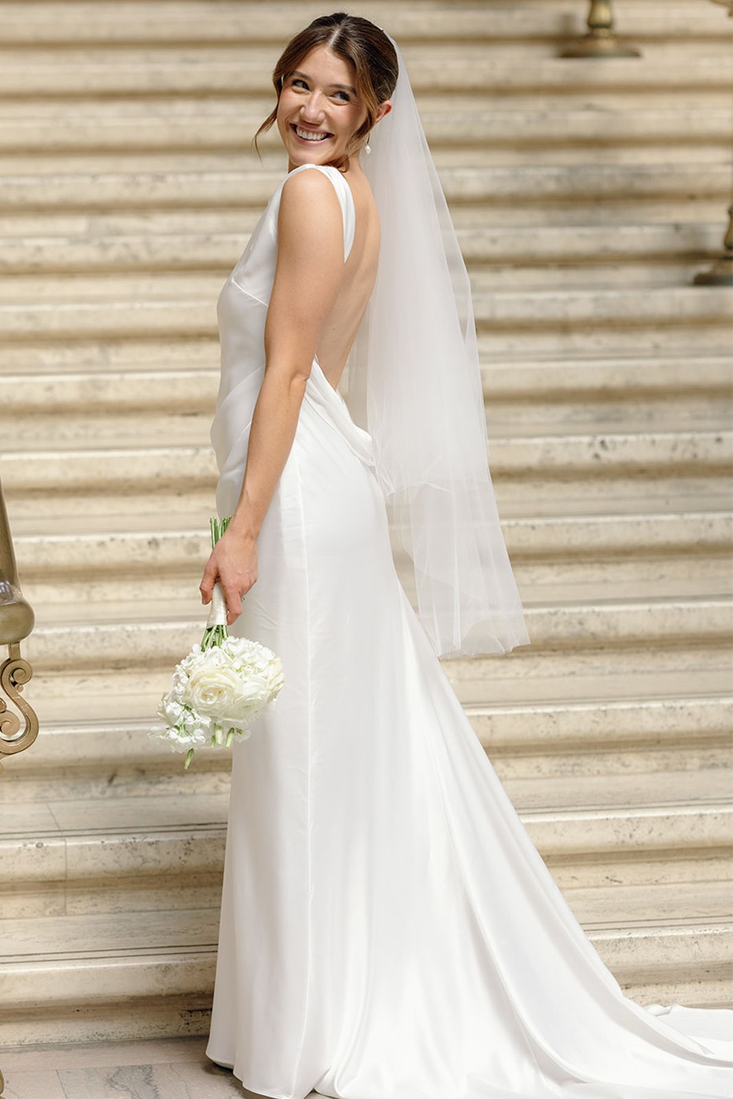 Bride in a white gown holding a bouquet, smiling on staircase.