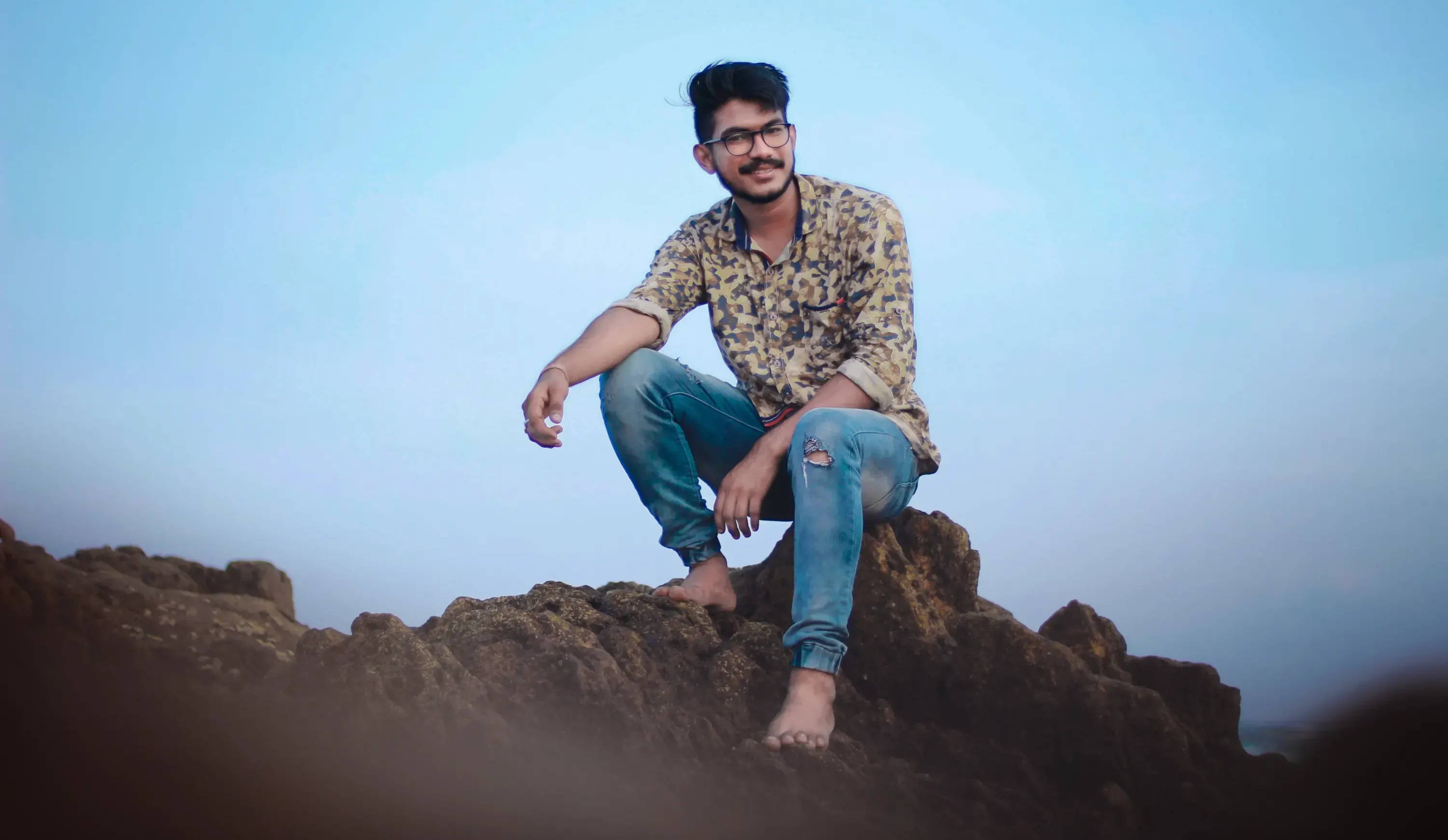 A man sits on a jagged rock in blue jeans and a button down shirt. Image by Vamsi Badireddi.
