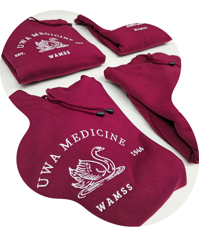 Folded maroon hoodies laid on white surface each embroidered with a white swan crest and UWA Medicine Est.1946 WAMSS text