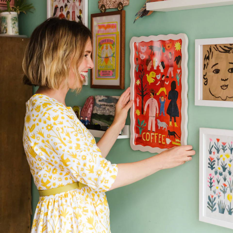 An image of Louise Lockhart wearing a dress with a bright yellow flower and bird print hanging a Printed Peanut picture on a wall. 
