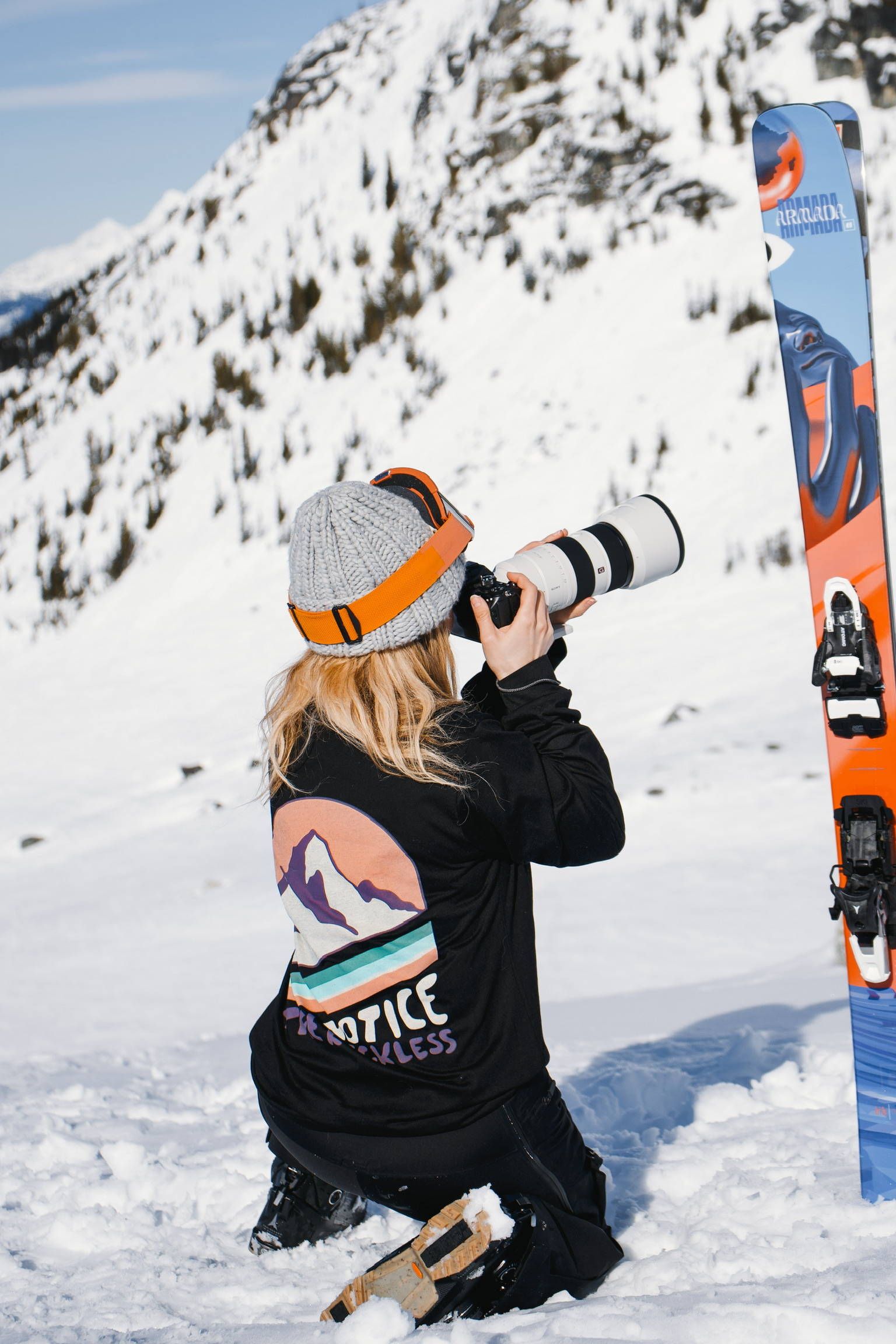 A woman kneeling down on the snow photographing the mountains with skis beside her