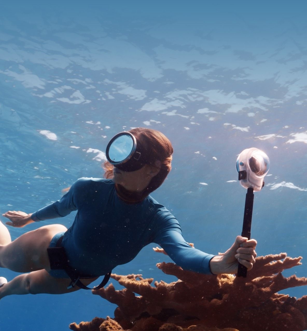 A person snorkeling near coral, holding a DJI Osmo 360 II camera with a waterproof case and selfie stick..