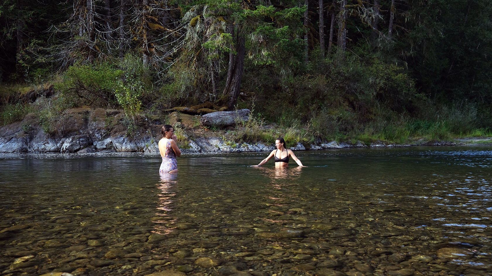 Two people wading in a clear river surrounded by trees.