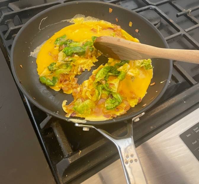 A top-view of scrambled eggs with greens and onions being made in a Misen Nonstick Pan. An unseen hand uses a wooden spoon to stir the eggs.