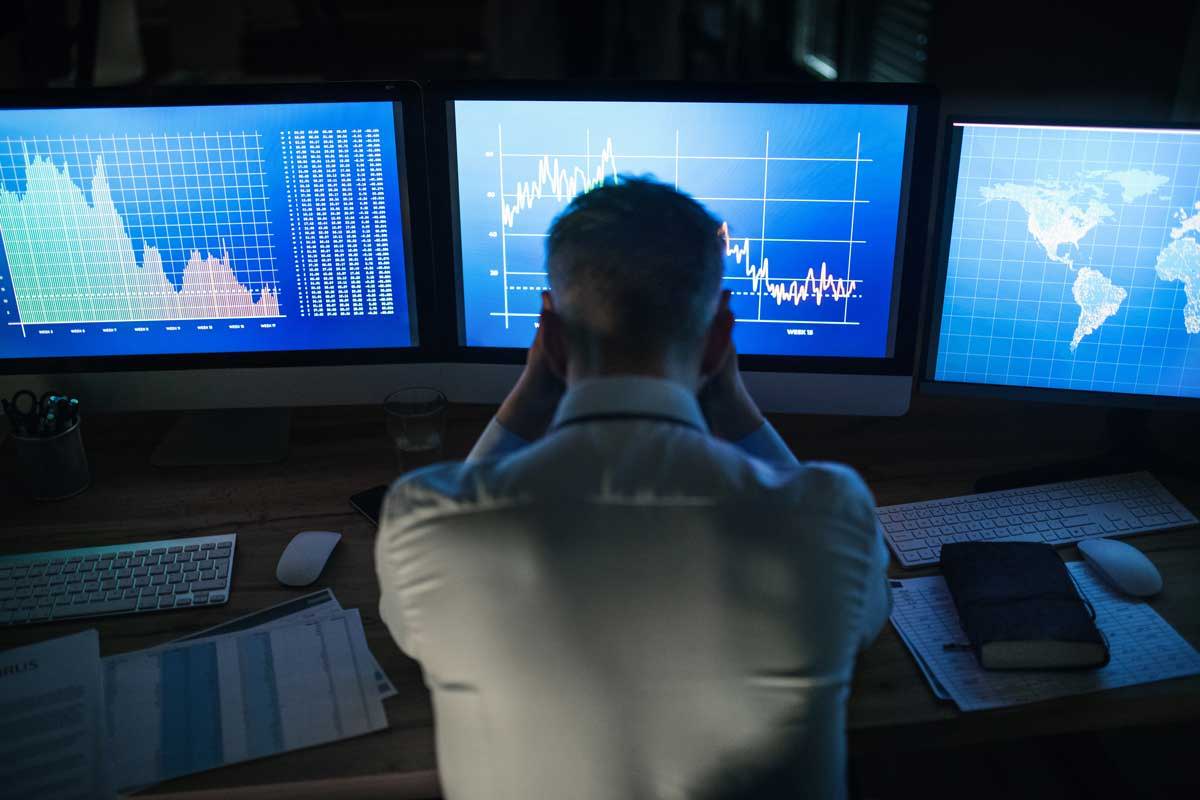 A man looking at three computer screens that show stock prices falling rapidly