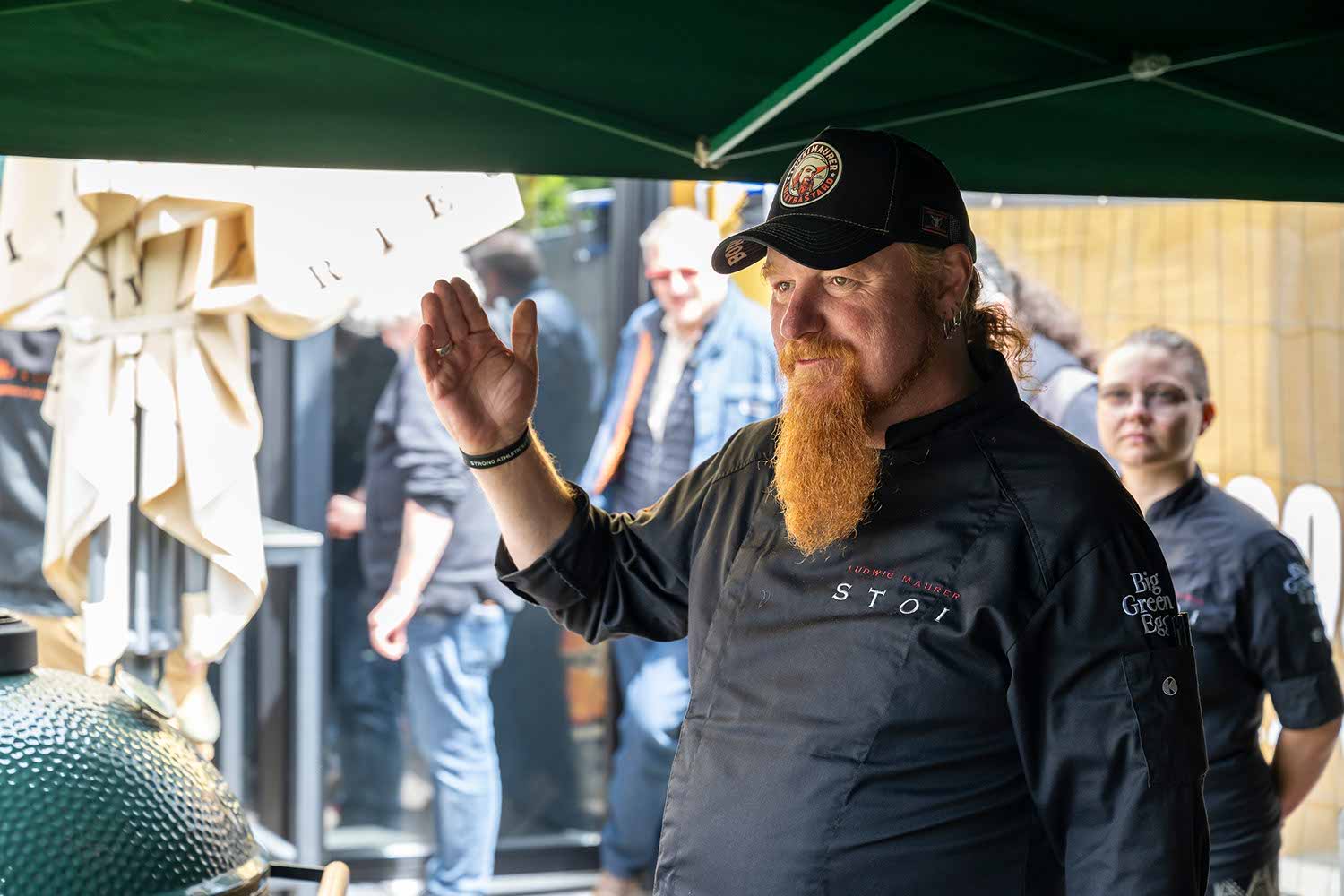 A chef with a beard gestures while cooking outdoors.