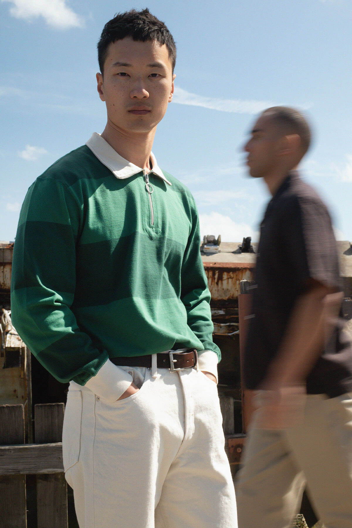 A person in a green shirt stands still, while another moves blurred in the background.
