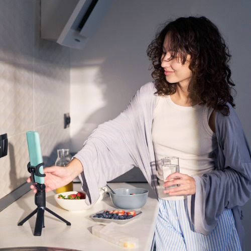 Woman holding a glass of water while creating content with a smartphone in her kitchen.