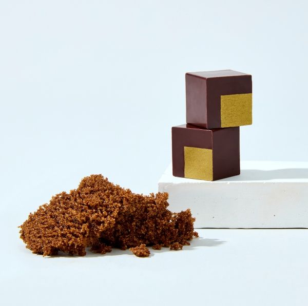 Two chocolate squares with gold accents next to brown sugar on a white surface.