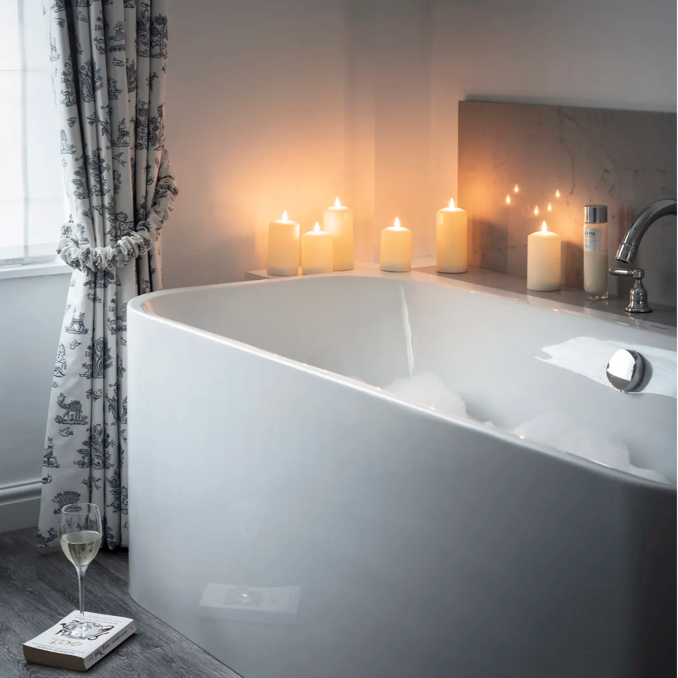 A bathroom setting with a bubble bath, LED candles and a glass of white wine.