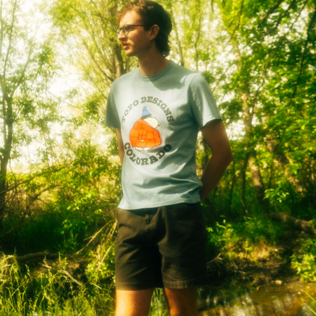 Person standing by a stream, wearing a t-shirt and shorts, surrounded by greenery.