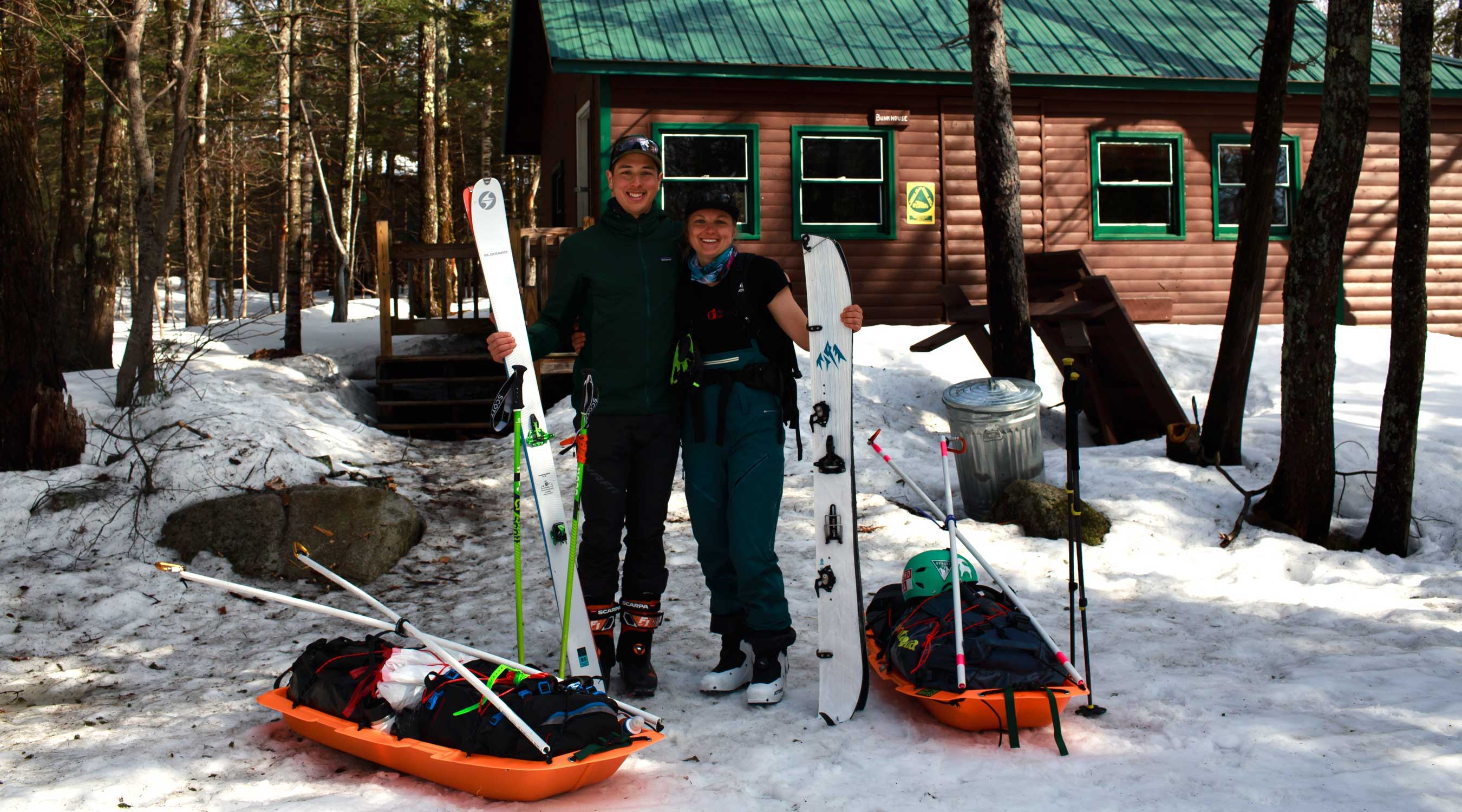 one skier beside one splitboarder pose with equipment in front of a cabin in the snow.
