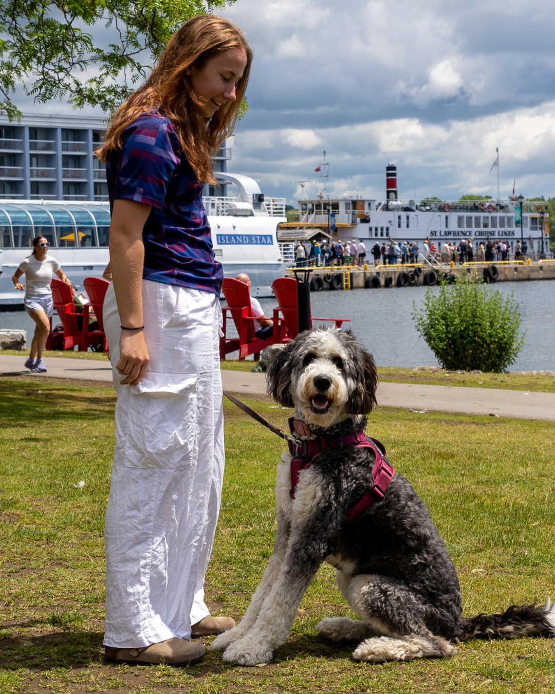 A woman standing next to a sitting dog by the water.