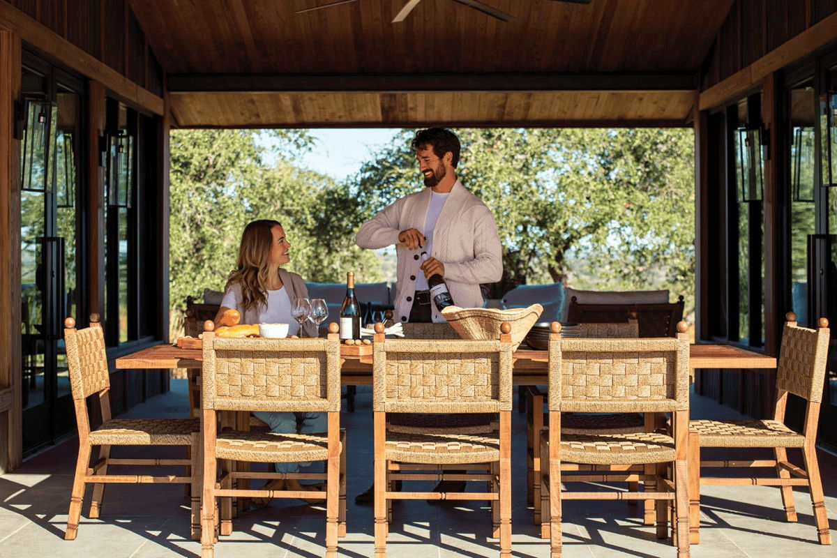 Couple at a long outdoor dining table with woven chairs under a wood-covered patio, set with wine glasses and dinnerware.