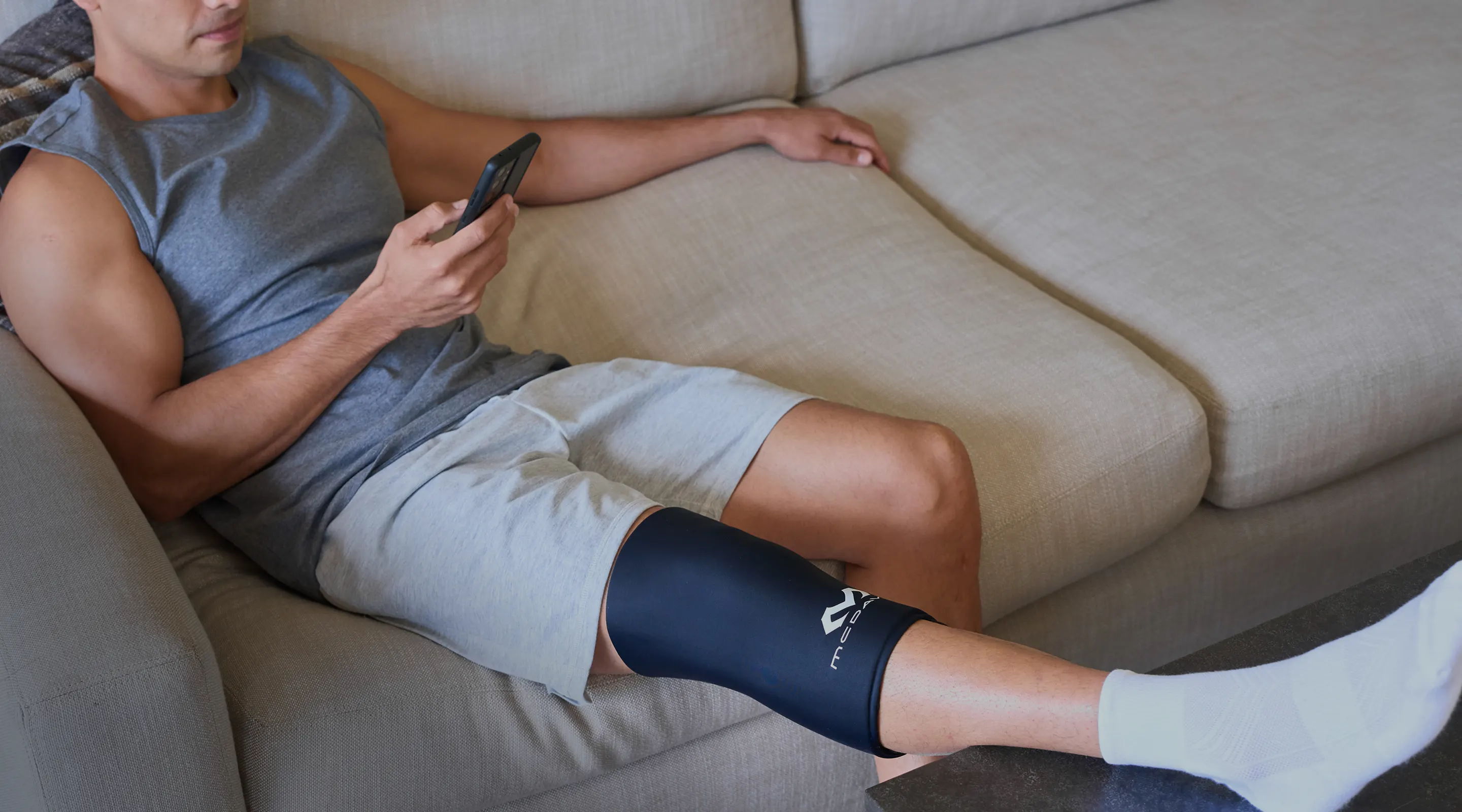 Person sitting on a couch, using a phone, wearing a ice recovery knee sleeve