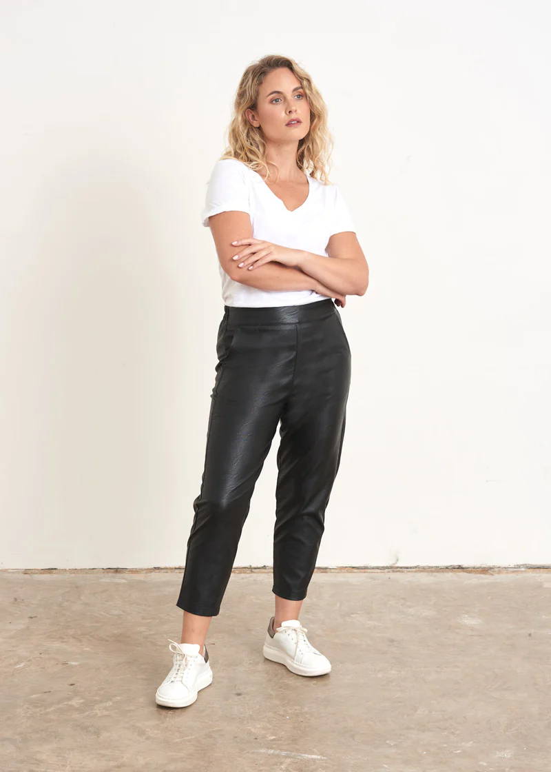 A model wearing a pair of faux leather black tapered trousers with a white top and white trainers