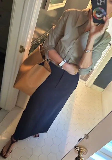 Person taking a mirror selfie, wearing a shirt and long skirt, holding a bag.