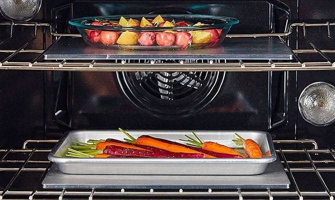 Two Misen Oven Steels cook vegetables on separate racks in one oven — one under a glass baking dish, the other under a metal sheet pan.