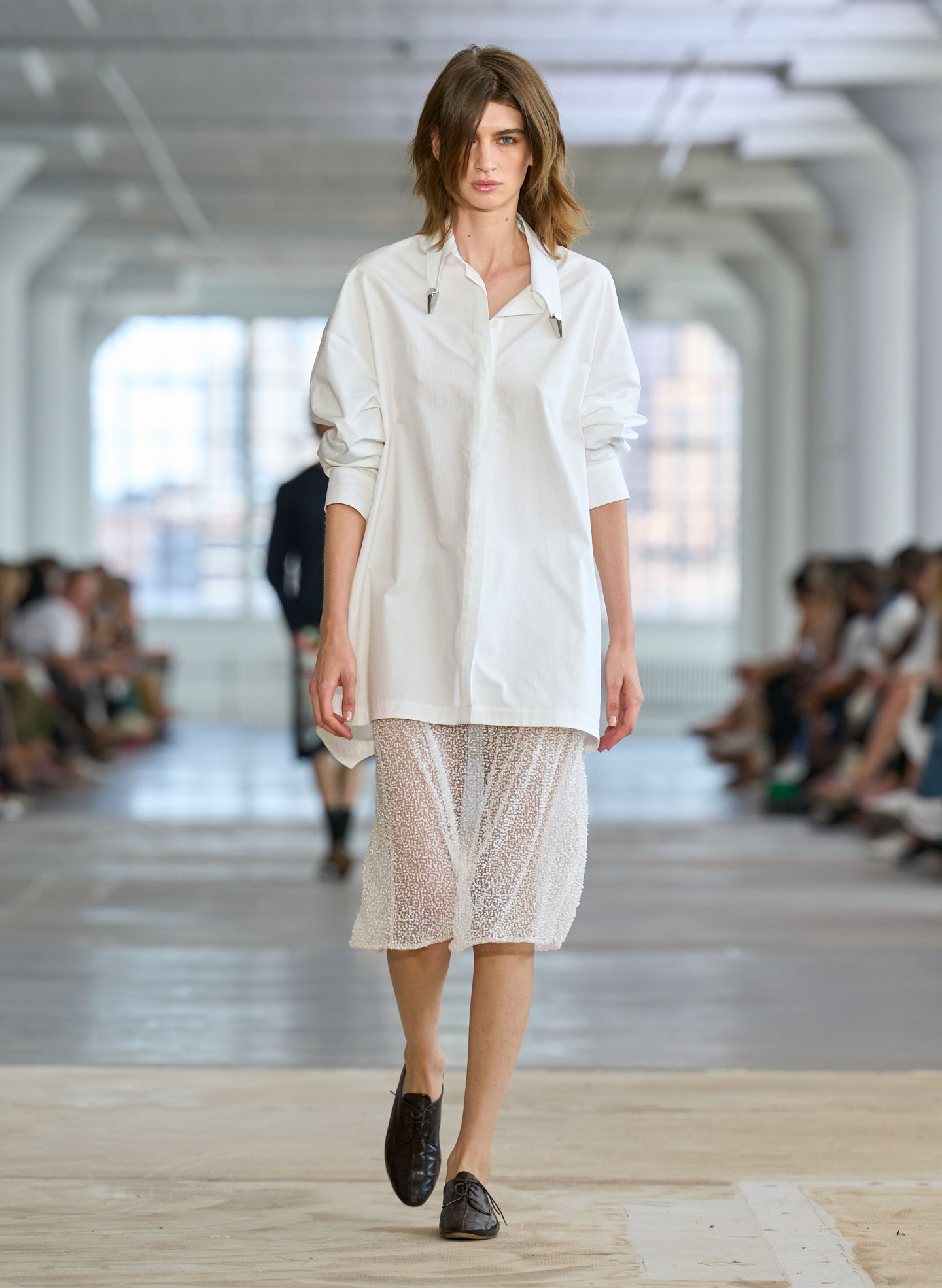 Model walking down runway in oversized white shirt and sheer skirt.