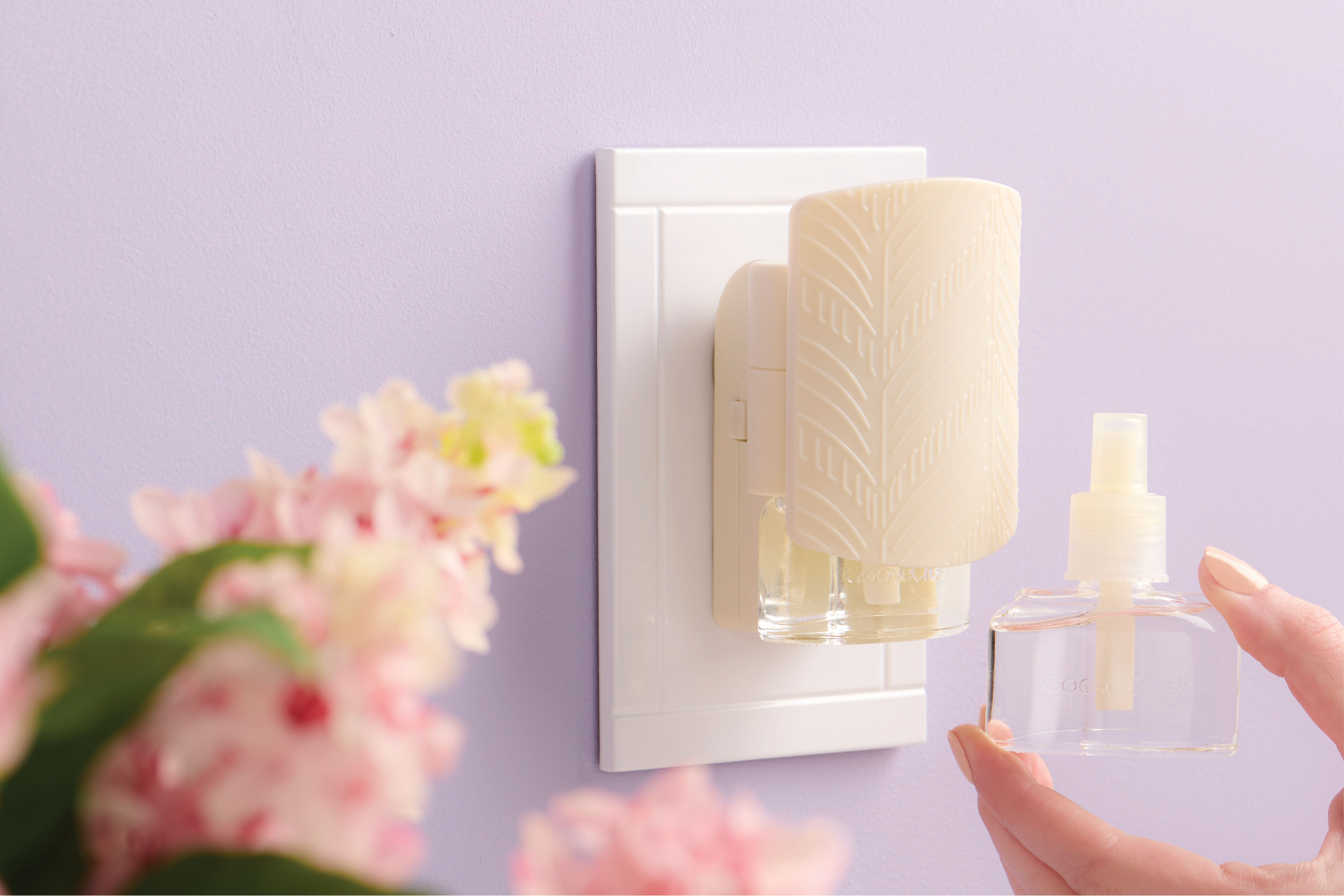 Hand holding a fragrance bottle near a wall-mounted diffuser and flowers.