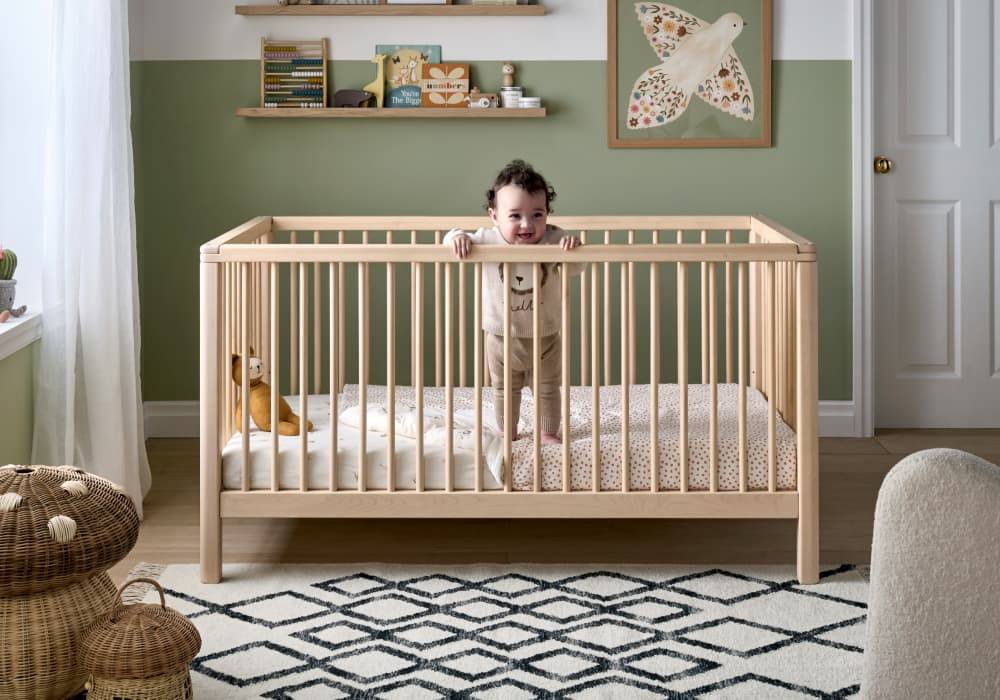A baby stands in their natural wooden crib, in a modern green nursery.