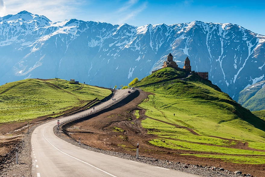 Curving road to Gergeti Trinity Church in Georgia, set against a backdrop of green hills and snowy Caucasus Mountains, highlighting the region's natural and historical beauty.