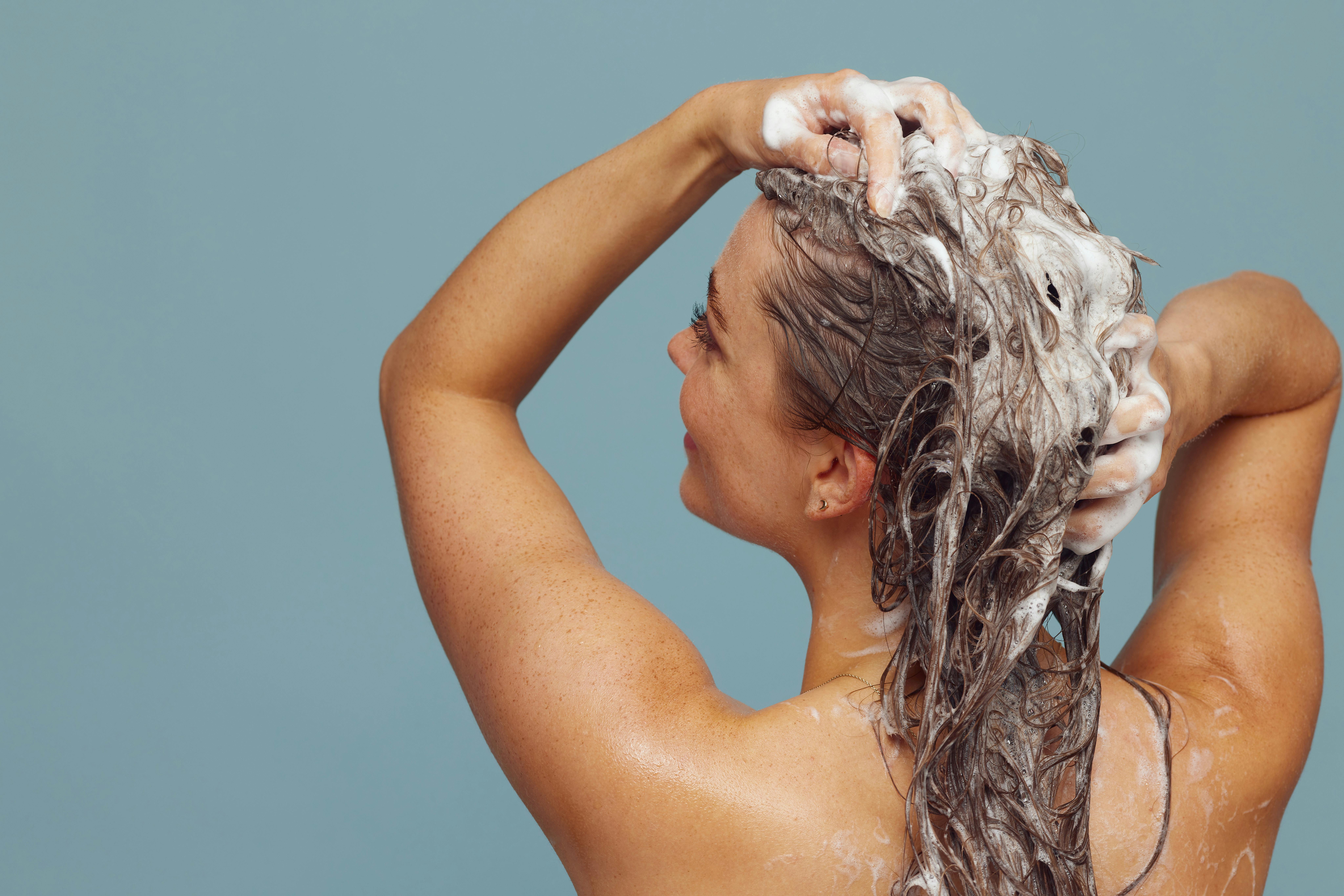 Woman applying shampoo to her hair against a blue background.