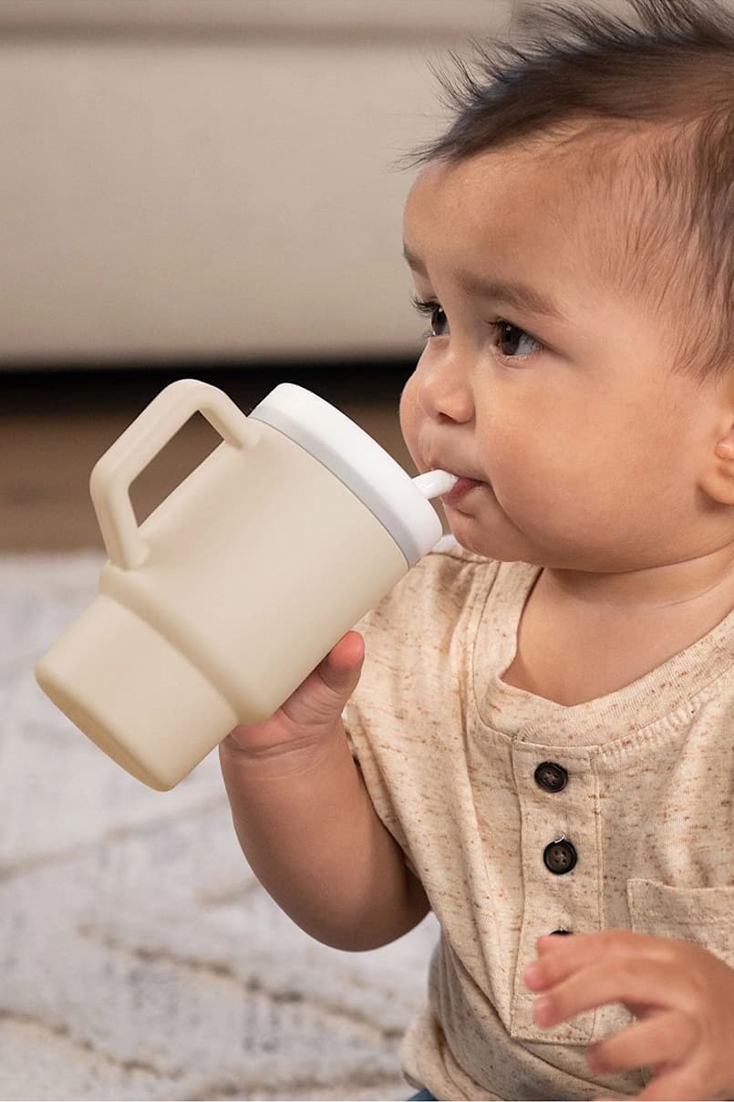 Baby drinking from a light-colored sippy cup while sitting on a rug.