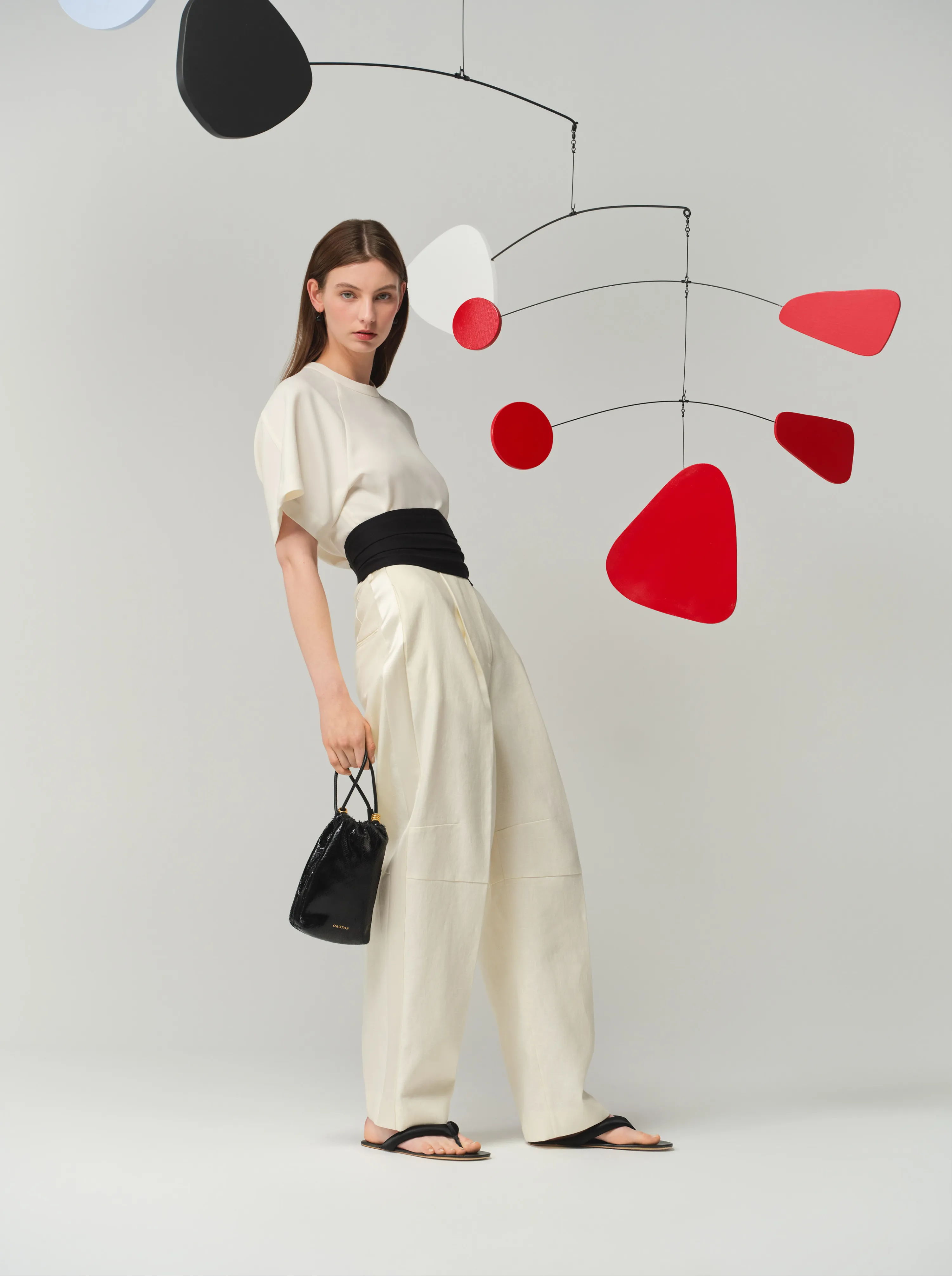 Model in cream outfit poses with a black bag and abstract mobile art.