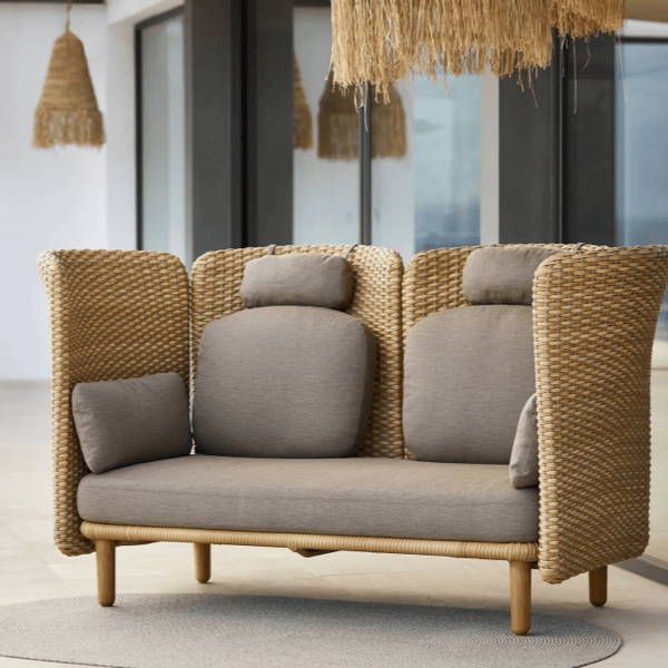 The Arch 2-Seater Outdoor Sofa featuring high arms, a woven natural rattan frame, and plush taupe cushions, positioned on a neutral patio with hanging woven light fixtures in the background. Perfect for cozy, modern outdoor retreats.
