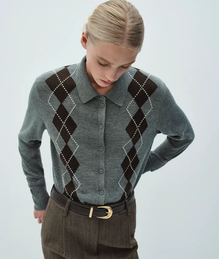 Woman wearing a gray argyle sweater and brown trousers, looking down.