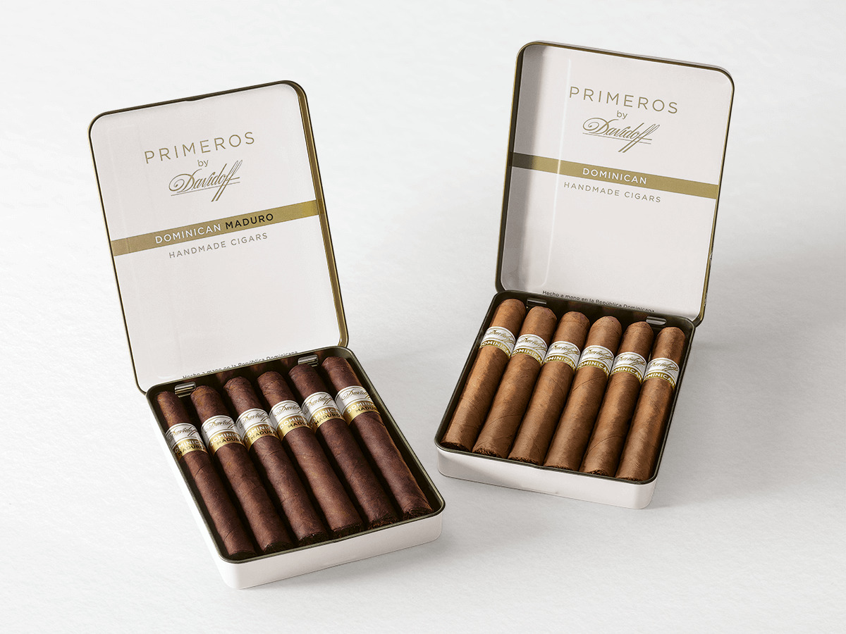 An opened tin of Primeros by Davidoff Dominican Maduro next to an opened tin of Primeros by Davidoff Dominican, both with petit panetela cigars inside. 