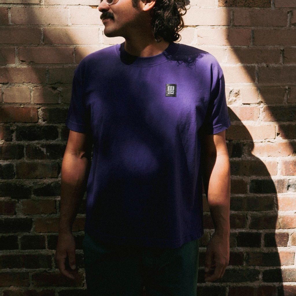 Person standing against a brick wall, wearing a purple shirt and looking sideways.