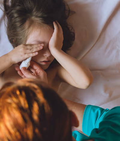 A girl lying in bed with her hands up to her face as mom leans over and gives her allergy eye drops to soothe the itching